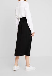 Gonna nera di media lunghezza con vita arricciata, abbinata a una camicia bianca con bottoni e sneakers bianche. Tessuto liscio con un design fluido.