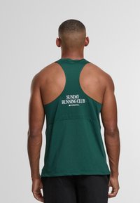 Muž v tmavě zeleném tričku bez rukávů s textem "Nedělní běžecký klub" na zadní straně, stojící před jednoduchým šedým pozadím.