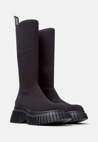 Botas negras hasta la rodilla hechas de material de punto acanalado, con una suela gruesa de goma estriada y una lengüeta para tirar en la parte superior.