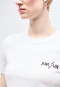 Άτομο που φοράει λευκό μπλουζάκι με ροζ αστραπή και τη λέξη "AMOUR" στο στήθος, εμφανίζοντας το λαιμό, τη γραμμή του σαγονιού και το σκουλαρίκι.