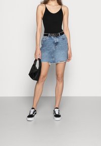 Jupe en denim avec ourlet effiloché, ceinture noire, body noir, et sac noir. Le mannequin porte des baskets noires et blanches, debout sur une surface gris clair.