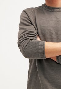 Pull en tricot gris avec des poignets et un col côtelés, arborant un motif texturé, porté avec les bras croisés.