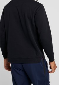 Schwarzer Pullover mit Rundhalsausschnitt, langen Ärmeln und gerippten Bündchen. Texturierte Stoffdetails entlang des Saums; ergänzt durch marineblaue Hose.