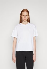 Weißes Baumwoll-T-Shirt mit kurzen Ärmeln, rundem Halsausschnitt und grünem gesticktem Logo auf der linken Brust. Dazu dunkle Hose.