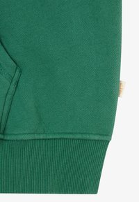 Tessuto di felpa verde con una texture liscia, polsini a costine e un'etichetta piccola con testo sulla cucitura laterale.