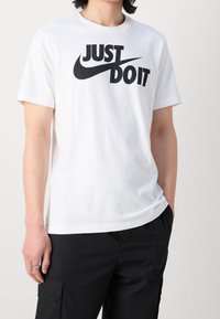 Vit bomulls-T-shirt med en stor svart "JUST DO IT"-logotyp med en swoosh-design. Korta ärmar och rund halsringning.