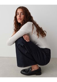 White long-sleeve top, dark blue wide-leg jeans, black loafers. Subject crouches with hands on knees, wavy hair framing the face.