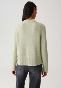 Woman seen from behind wearing a light green fuzzy sweater and dark gray jeans against a plain background.