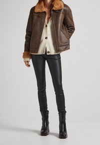 Chaqueta de cuero marrón con cuello de borreguito, cárdigan crema debajo, pantalones de cuero negros y botas negras de tobillo con tacón grueso.