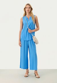 Top azzurro senza maniche con bottoni, abbinato a pantaloni blu a gamba larga. La modella porta una borsa a tracolla intrecciata beige e indossa sandali.