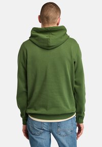 Felpa verde con cappuccio, dalla texture liscia, polsini e vita a costine. Presenta un cappuccio ampio e dettagli sulle cuciture posteriori. Abbinata a jeans blu.