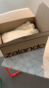Caja de cartón con la marca Zalando, que contiene una sudadera color crema doblada, colocada sobre una mesa de mármol con patas rojas.