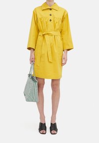 Abito di cotone giallo con colletto, tre bottoni e vita legata. Abbinato a una borsa a righe verdi e bianche e sandali neri con tacco.