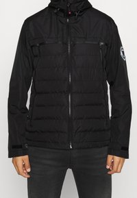 Mand iført en sort zip-up puffjakke med hætte og et patch på venstre ærme, parret med mørke bukser mod en ensfarvet baggrund.