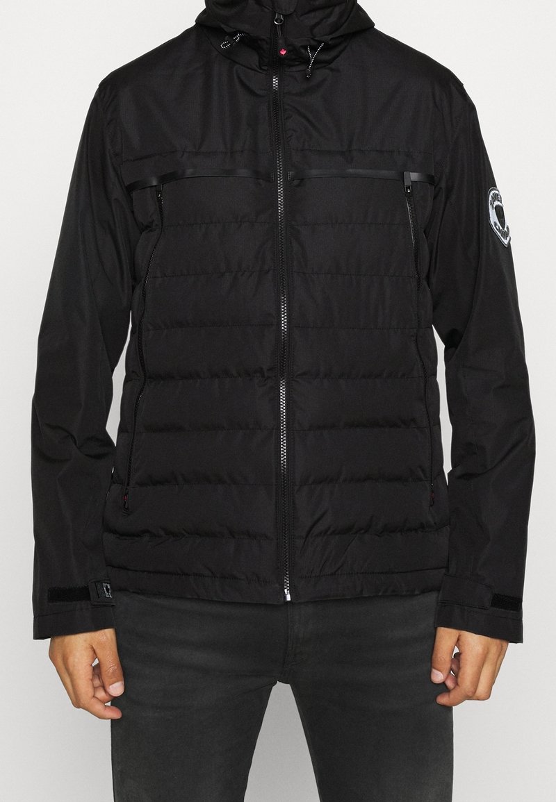 Mand iført en sort zip-up puffjakke med hætte og et patch på venstre ærme, parret med mørke bukser mod en ensfarvet baggrund.