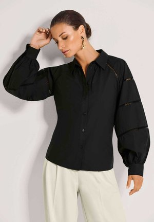 Zwart overhemd met knopen, pofmouwen, kanten details op de armen en een klassieke kraag; gedragen met lichtgekleurde broek.