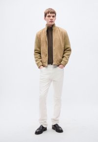 Μπεζ μπουφάν bomber από σουέτ με μανίκια από πλεκτό ύφασμα, καφέ ζιβάγκο, λευκό παντελόνι και μαύρα παπούτσια. Απλός, μοντέρνος σχεδιασμός και ουδέτερα χρώματα.