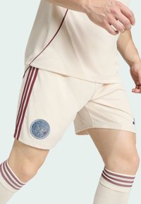 Mężczyzna w kremowych szortach sportowych i skarpetach w bordowe paski, stojący z nogami rozstawionymi i rękami lekko uniesionymi na jasnym tle.