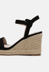 Sandale compensée en daim noir avec bride à la cheville, semelle plateforme en jute tressé et boucle réglable sur fond blanc.