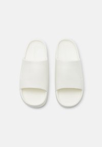 Hvite slip-on sandaler med en glatt, matt tekstur; åpen tå og bred rem over foten. Teksturert såle for bedre grep.