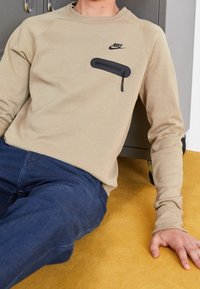 Osoba ubrana w beżową bluzę Nike z czarną kieszenią na suwak, siedząca na żółtej podłodze, w ciemnoniebieskich spodniach.