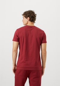 Tricou din bumbac maron, cu mâneci scurte, guler rotund și detalii subtile cu logo pe spate. Materialul are o textură netedă.