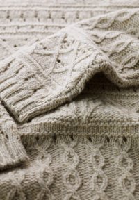 Sweter robiony na drutach w jasnym beżowym kolorze z wełny, z teksturowanymi wzorami i splotami, z podwiniętymi krawędziami i miękkim, puszystym wyglądem.