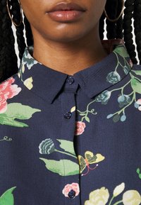 Camicia floreale con sfondo blu navy, caratterizzata da motivi di fiori colorati, colletto button-down e tessuto testurizzato. Due bottoni blu al collo.