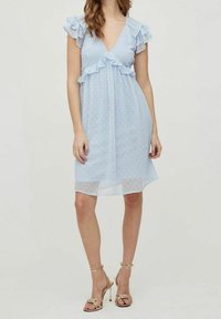 Ljusblå klänning med rynkade axelband, V-ringning och prickigt mönster, med ett flödande, genomskinligt överdrag och en åtsittande bodice.