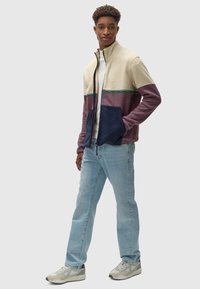 Chaqueta de forro polar con cremallera que presenta bloques de color en crema, burdeos y navy, con una superficie texturizada y costuras en verde contrastante. Combinada con jeans claros.