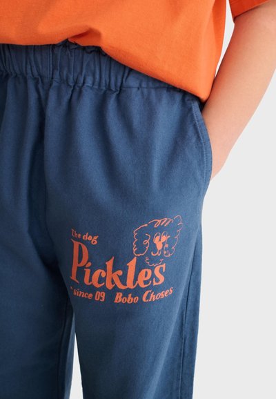 Child wearing blue elastic-waist pants with orange dog illustration and text "Pickles since 09 Bobo Choses" on thigh, hand in pocket.
