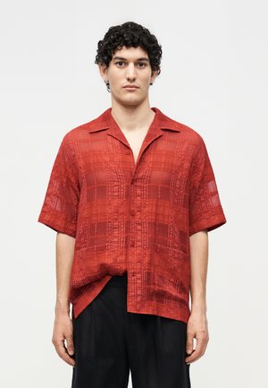 Homme aux cheveux bouclés foncés, portant une chemise rouge texturée à manches courtes et un pantalon noir, debout devant un fond uni clair.