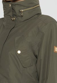 Veste vert olive à col montant avec une poche poitrine zippée, un rabat à bouton-pression et un petit patch de marque rectangulaire sur la manche.