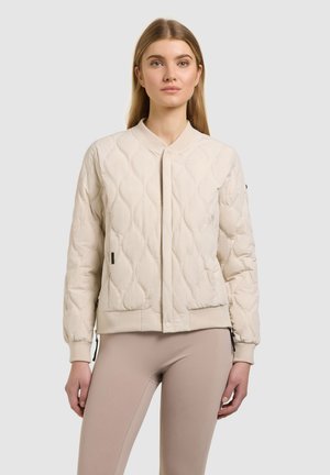 Młoda kobieta w beżowej pikowanej bomberce i jasnobrązowych dopasowanych spodniach, stojąca na tle jednolitego jasnoszarego tła.