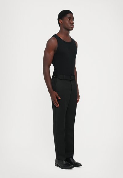 Un top negru fără mâneci și pantaloni negri ajustați, purtați cu un curea și pantofi de ceremonie negri. Texturi netede; design ajustat. Poziție în picioare pe un fundal alb.