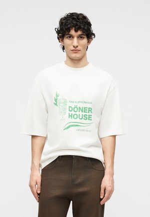 Νεαρός με σγουρά μαλλιά φοράει λευκό φαρδύ T-shirt με πράσινο γράφημα "Döner House" και καφέ παντελόνι, στέκεται μπροστά σε απλό φόντο.