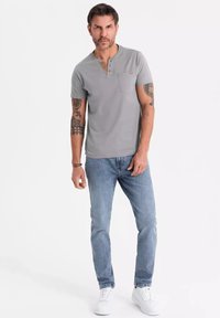 Camisa gris de manga corta con un pequeño bolsillo en el pecho y tapeta de tres botones, combinada con jeans de mezclilla azul claro y zapatillas blancas.