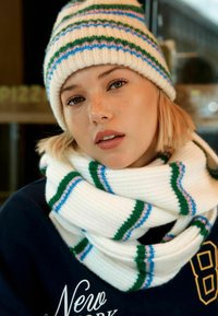 Kootud valge beanie roheliste, siniste ja roosade triipudega. Paks valge sall sarnaste triipudega, kantud musta sweatshirt'i all, millel on tekst.