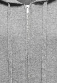 Sweat à capuche gris zippé en tissu doux, avec une fermeture éclair à l'avant, des coutures texturées, et une capuche avec cordons de serrage.