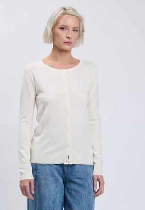 Lyshåret kvinde iført en hvid knappet cardigan og blå jeans står mod en ensfarvet lys baggrund.