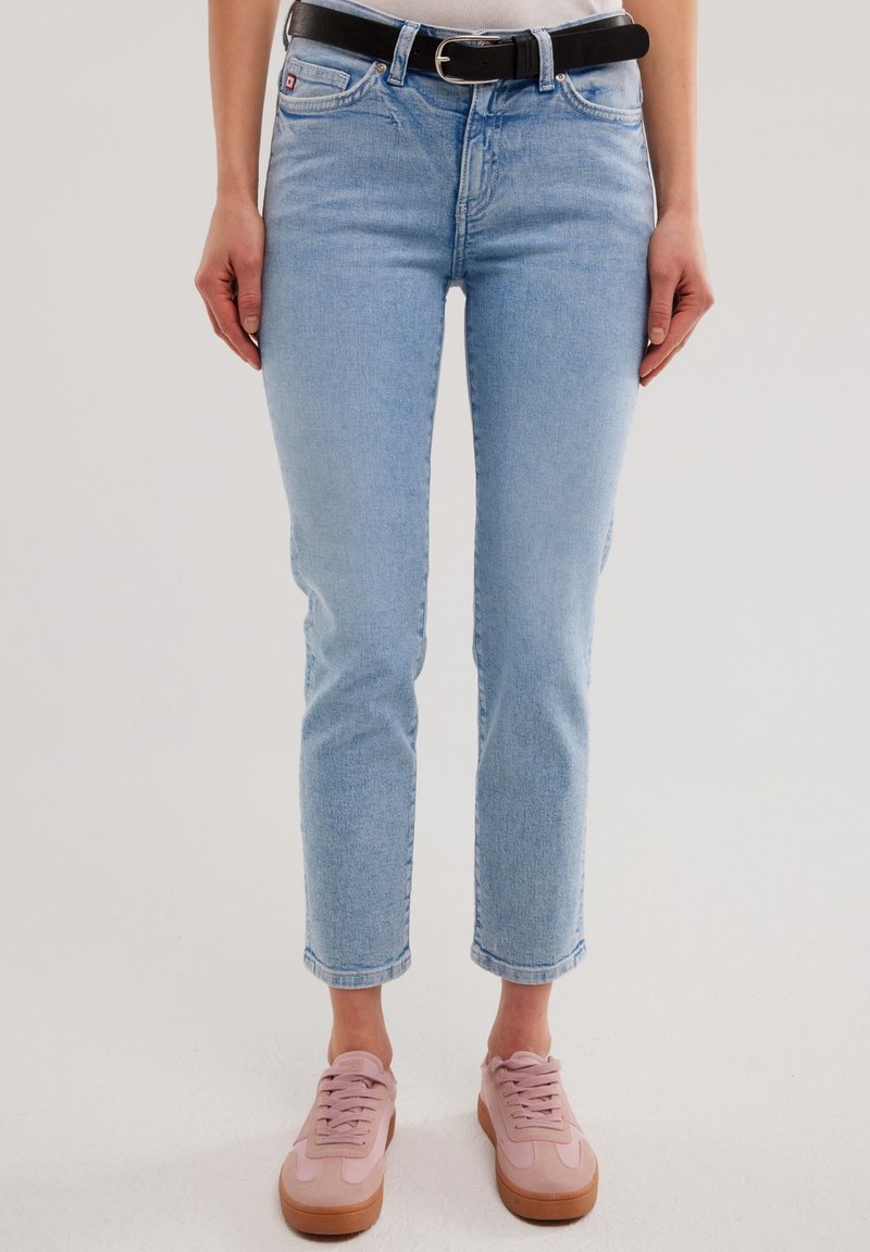 Personne portant un jean bleu clair, une ceinture noire, un haut blanc et des baskets roses, debout devant un fond clair et uni.
