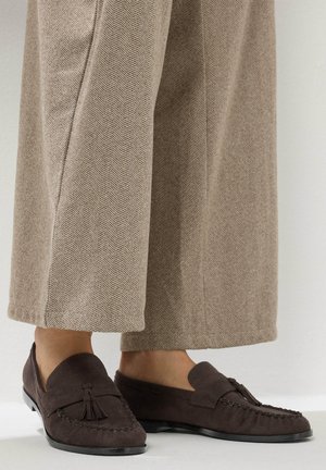 Bruine suède loafers met kwastjes, gedragen met beige wijde broekspijpen in visgraatmotief, waarbij de onderbenen en voeten zichtbaar zijn, staand op een witte vloer.