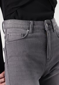 Pilkos džinsinės kelnės su prigludusiu dizainu, turinčios kišenes ir užtrauktuką. Audinys turi šiek tiek tekstūros ir subtilių siuvimo detalių.