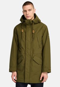 Olivgrön parkajacka med huva, utrustad med två framfickor, snörningsdetaljer och en texturerad tygfinish.