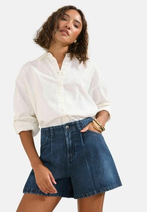 Frau trägt ein lockeres weißes Hemd mit Knöpfen und hochgekrempelten Ärmeln sowie hoch taillierte dunkle Jeansshorts, accessoriert mit goldenen Armreifen.