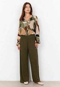 Flerfärgad blus med geometriska mönster i nyanser av grön, beige och brun; kombineras med texturerade olivgröna vida byxor och beiga lägenheter.