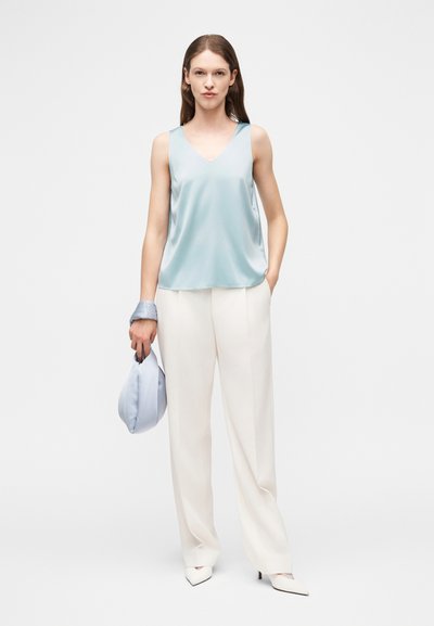 Γυναίκα που φοράει ανοιχτό γαλάζιο αμάνικο τοπ και άσπρο παντελόνι με φαρδιά πόδια, κρατώντας τσάντα σε ίδιο ανοιχτό γαλάζιο χρώμα με μαντήλι τυλιγμένο στον καρπό.