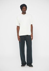 Λευκή κοντομάνικη μπλούζα με μαύρα γράμματα, σε συνδυασμό με φαρδιά σκούρα τζιν. Το μοντέλο φοράει ένα σκούρο πλεκτό καπέλο και μαύρα παπούτσια.