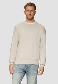 Man in een beige ronde hals trui over een wit shirt en lichtblauwe gescheurde jeans, staande tegen een effen lichte achtergrond.
