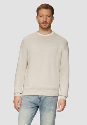 Man in een beige ronde hals trui over een wit shirt en lichtblauwe gescheurde jeans, staande tegen een effen lichte achtergrond.
